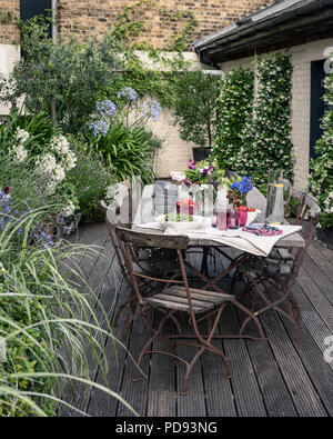 Alte französische Bistro Stühle um langen Holztisch auf Garten Dachterrasse mit Belag. Agapanthus und Jasmin blüht im Hintergrund Stockfoto