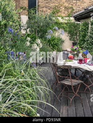 Alte französische Bistro Stühle um langen Holztisch auf Garten Dachterrasse mit Belag. Agapanthus und Jasmin blüht im Hintergrund Stockfoto