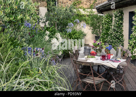 Alte französische Bistro Stühle um langen Holztisch auf Garten Dachterrasse mit Belag. Agapanthus und Jasmin blüht im Hintergrund Stockfoto