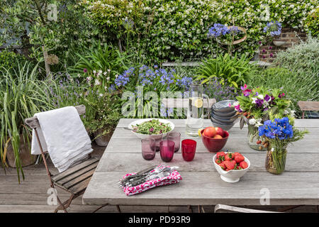 Rustikalen Holztisch auf Garten Dachterrasse für ein leichtes Mittagessen im Freien. Agapanthus und Jasmin im Hintergrund blühen Stockfoto