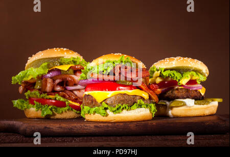 Drei verschiedene leckere Burger mit Rindfleisch, Tomaten, Käse, Gurken, Zwiebeln und Salat auf Holztisch und braunen Hintergrund mit Kopie Raum Stockfoto