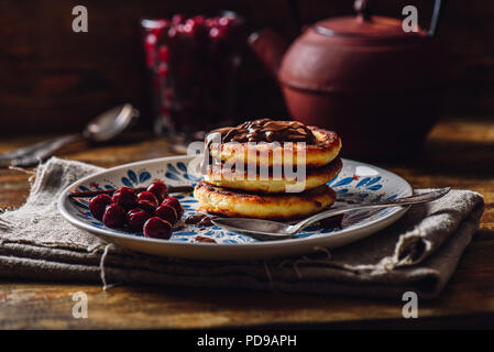 Frühstück mit Quark Pfannkuchen mit Schokolade einfügen und gefrorene Kirschen. Teekanne mit Löffeln und Glas von Beeren für den Hintergrund. Stockfoto