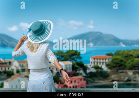 Assos im Morgenlicht. Junge touristische Frau trägt blaue Sonnenhut und weiße Kleidung bewundernden Blick auf bunte ruhige Dorf. Genießen Sommer Tag Reisen Urlaub auf Kefalonia, Griechenland Stockfoto