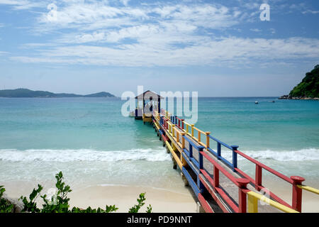 Bunte Beton und Holzsteg, über einem weißen Sandstrand eine klare blaue Meer, Felsen und Bäume auf beiden Seiten der Himmel mit Whi blau Stockfoto