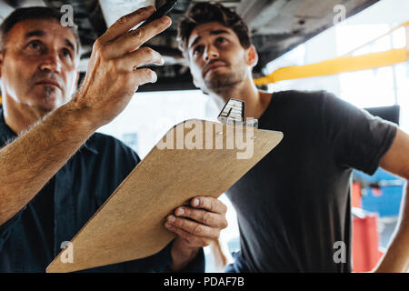 Zwei erfahrene Mechaniker stehen unter einem Auto und untersuchen das Problem. Mechanik, Auto Inspektion mit Zwischenablage. Stockfoto