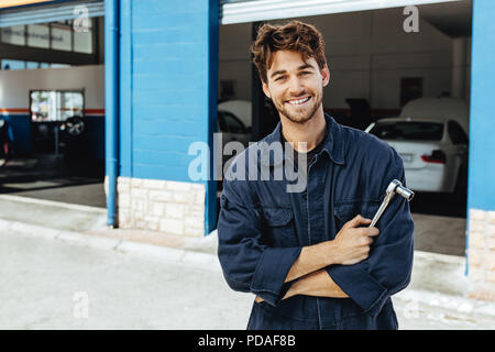 Professionelle Automechaniker in Uniform mit dem Schraubenschlüssel. Junge Mechaniker stehen mit verschränkten Armen außerhalb seiner Garage. Stockfoto