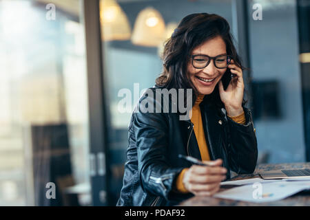 In casuals im Büro sitzen auf einem Tisch reden über Mobiltelefon Geschäftsfrau. Frau Unternehmer ihr Unternehmen verwalten. Stockfoto