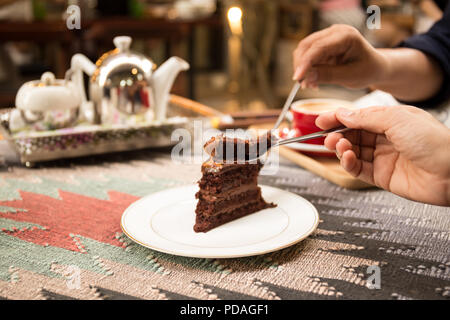 Woman's Hand schneiden ein Stück Schokoladenkuchen mit Kaffee Topf im Hintergrund. Stockfoto