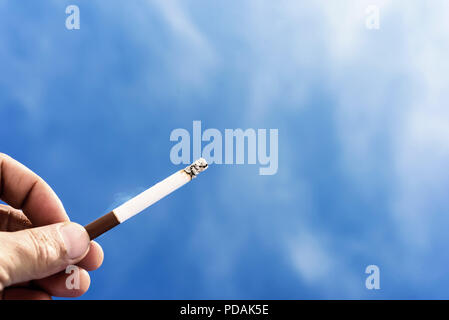 Einzelne männliche Hand mit einer Zigarette vor blauem Himmel Hintergrund mit Fetzen von giftigen Rauch aus dem Ende der Cig. Copyspace Bereich für ni Stockfoto