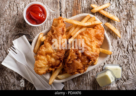 Lecker Fisch und Chips in einem nehmen Sie Behälter mit Ketchup, Sauce Tartar, Papierservietten und einen Kunststoff Gabel auf einem rustikalen Holztisch. Stockfoto