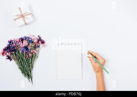 Weibliche hand mit Bleistift in der Nähe von offenen Notepad, Bündel wilder Blumen und kleine Geschenkbox auf Weiß, Ansicht von oben. Flach feier Vorbereitung Mock-up von wo Stockfoto