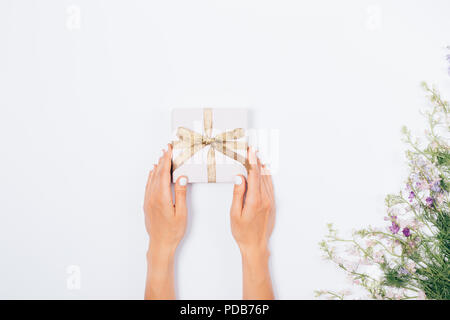 Woman's Hände, die kleine Schachtel in der Nähe von dekorativen Blumenmotiv auf weißem Hintergrund, Ansicht von oben. Flach Mädchen, die eine Neben Bouquet von Co vorhanden Stockfoto