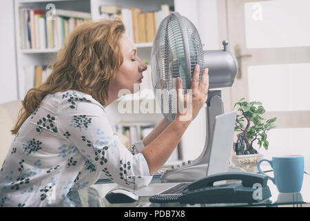 Frau leidet unter der Hitze, während der Arbeit im Büro und versucht durch den Lüfter zu kühlen Stockfoto