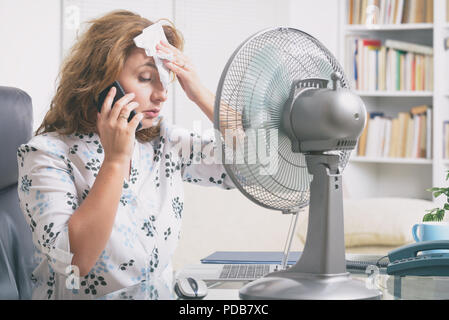 Frau leidet unter der Hitze, während der Arbeit im Büro und versucht durch den Lüfter zu kühlen Stockfoto