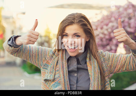 Helle junge Frau auf der Suche super halten Daumen hoch in Genehmigung und lächelnd in die Kamera im Freien begeistert Stockfoto