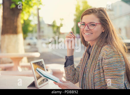 Hübsche stilvolle hipster Frau in Gläsern sitzen auf der Straße mit Tablet und Notepad Studien in und lächelnd an Kamera Stockfoto