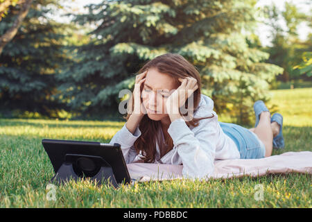 Porträt der jungen weißen Kaukasischen liegende Frau im Park auf Gras Ansehen digitalen Tablet. Mädchen Schüler besorgt Gefühl hervor. Starke Emotion, lustig f Stockfoto