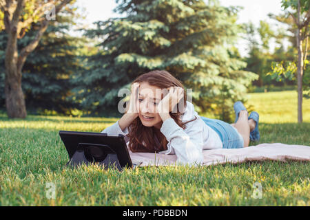 Porträt der jungen weißen Kaukasischen liegende Frau im Park auf Gras Ansehen digitalen Tablet. Mädchen Schüler besorgt Gefühl hervor. Starke Emotion, lustig f Stockfoto