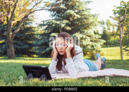 Porträt der jungen weißen Kaukasischen liegende Frau im Park auf Gras Ansehen digitalen Tablet. Mädchen Schüler besorgt Gefühl hervor. Starke Emotion, lustig f Stockfoto