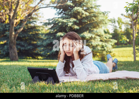 Porträt der jungen weißen Kaukasischen liegende Frau im Park auf Gras Ansehen digitalen Tablet. Mädchen Schüler besorgt Gefühl hervor. Starke Emotion, lustig f Stockfoto