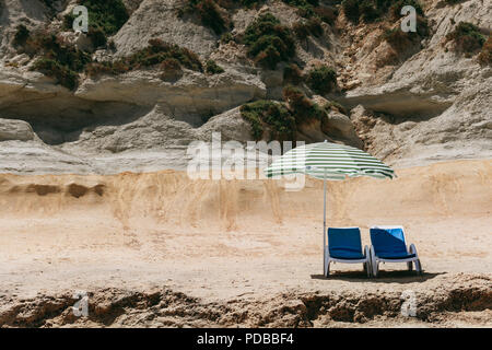 Zwei blaue Liegestühle mit Sonnenschirm am Strand mit grünen Streifen in einem einsamen Strand. Stockfoto