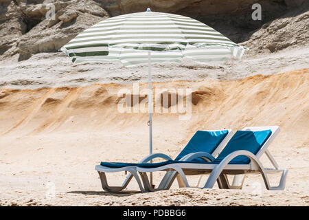 Zwei blaue Liegestühle mit Sonnenschirm am Strand mit grünen Streifen in einem einsamen Strand. Stockfoto