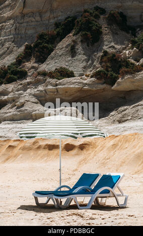 Zwei blaue Liegestühle mit Sonnenschirm am Strand mit grünen Streifen in einem einsamen Strand. Stockfoto