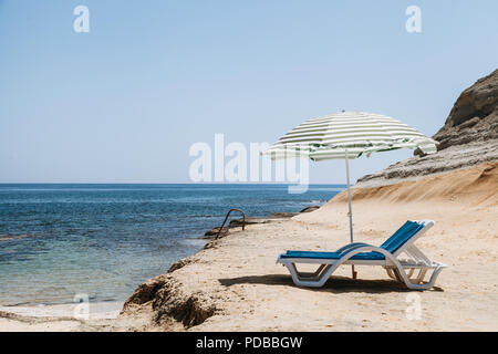 Zwei blaue Liegestühle mit Sonnenschirm am Strand mit grünen Streifen in einem einsamen Strand. Stockfoto