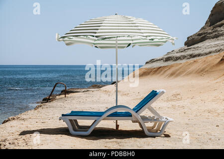 Zwei blaue Liegestühle mit Sonnenschirm am Strand mit grünen Streifen in einem einsamen Strand. Stockfoto