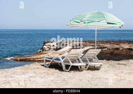 Zwei blaue Liegestühle mit Sonnenschirm am Strand mit grünen Streifen in einem einsamen Strand. Stockfoto