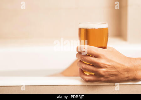 Nahaufnahme eines jungen kaukasischen Mann trinkt ein erfrischendes Bier, während sich bei einem Bad in der Badewanne von einem beigefarbenen Badezimmer Stockfoto