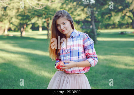 Portrait von schönen jungen kaukasischen Frau mit langen roten Haaren in Plaid Shirt und rosa Tutu Tüllrock, im Park Wiese am Sommer, Sonnenuntergang, in suchen Stockfoto