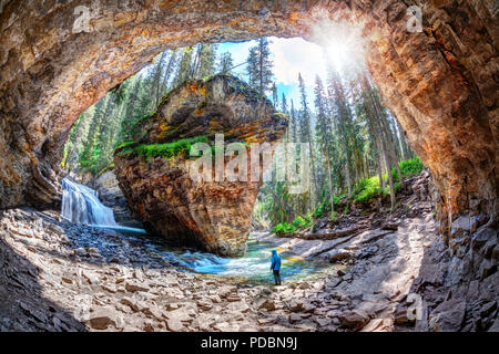 Wanderer steht in Ehrfurcht vor Wasserfall und Kalksteinschichten in eine versteckte Höhle in der Johnston Canyon im Banff National Park, mit Sun platzen durch die üppigen Stockfoto