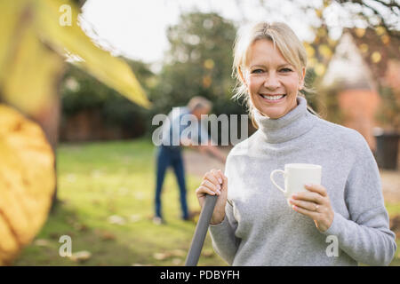 Porträt Lächeln, zuversichtlich, reife Frau trinkt Kaffee und Harken Blätter im Herbst im Hinterhof Stockfoto