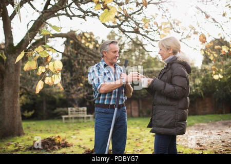 Reifes Paar Kaffee trinken und Harken Blätter im Herbst im sonnigen Hinterhof Stockfoto