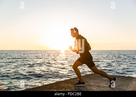 Sportliche erwachsene Mann in Kopfhörern tun Bein eine hockt während des Trainings am Meer Stockfoto