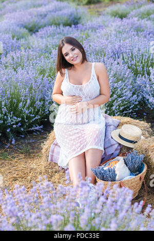 Lächelnd schwangere Frau sitzt auf der Heu Ballen in violett Lavendel Feld berühren und Bauch Stockfoto