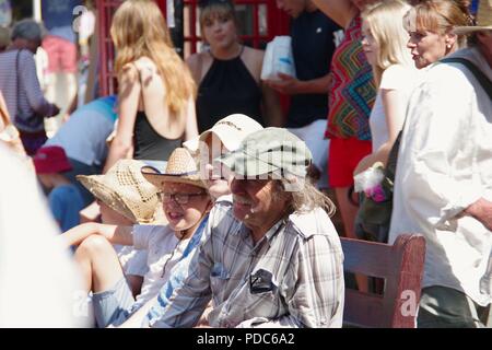 Großeltern und Enkel auf einer Holzbank Inmitten einer Menschenmenge beobachten Veranstaltung in Sidmouth Folk Festival. East Devon, Großbritannien. August, 2018. Stockfoto