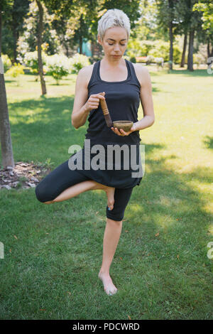 Frau Yoga in Baum und Sound mit tibetischen Klangschalen in Park Stockfoto