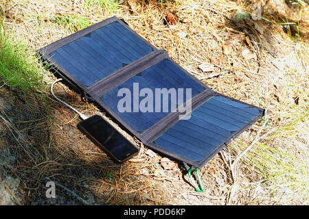 Das faltbare Solarmodul liegt auf der Oberfläche des Waldbodens und Gebühren das Smartphone. Komfort für Reisen und lange Spaziergänge vom traditio Stockfoto