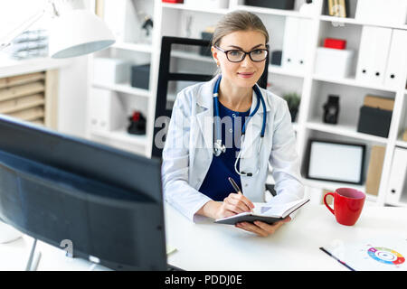 Ein junges Mädchen in einem weißen Gewand sitzt an einem Tisch im Büro und hält einen Stift und ein Notebook in der Hand. Ein Stethoskop um den Hals hängt. Stockfoto