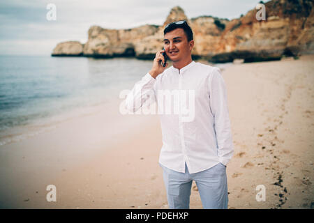 Junge Mann ist mit seinem Handy am Strand im Sonnenuntergang Stockfoto