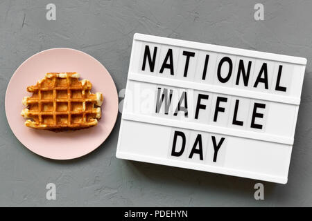 "Nationale Waffel Tag' Wort auf Lightbox, traditionelle belgische Waffel auf rosa Platte über konkrete Oberfläche, Ansicht von oben. Von oben und Overhead. Stockfoto