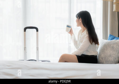 Entspannen Sie nach einem harten Arbeitstag. Schöne Asiatische junge lächelnde Geschäftsfrau im Anzug Kaffee trinken und weg schauen beim Sitzen auf einem Sofa im Hotel r Stockfoto