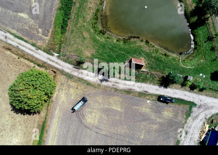 Vertikale Luftaufnahme über einen Weg neben einem Abgeernteten Feld und eine Wiese mit einem kleinen Stall vor einem Teich zum Angeln. Stockfoto