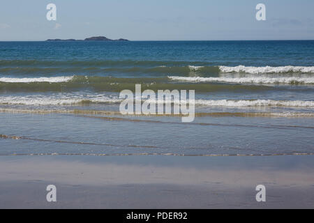 Ein Blick auf das Meer auf einem Waliser Strand mit sanften Wellen ans Ufer und eine Insel in der Ferne auf einem sonnigen klaren Tag Stockfoto