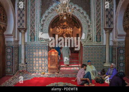 Fes, Marokko - Mai 11, 2013: Die Menschen sitzen durch den Eingang zu einer Moschee, mit kunstvollen Mosaik und Arabesken Schnitzereien verziert Stockfoto