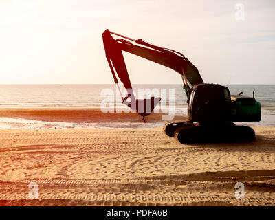 Bagger arbeiten am Strand mit Blick auf den Sonnenuntergang. Silhouette von Bagger und Reifenspur auf Sand mit Sonnenlicht am Abend auf Marine backgr Stockfoto