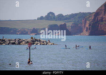 Menschen Abkühlung und Paddle Boarding in das Meer bei Sidmouth. Lardam Bay Sea Stacks und roten Felsen in der Ferne. East Devon, Großbritannien. August, 2018. Stockfoto