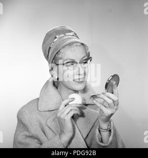 1950 Make-up. Eine junge Frau schaut sich in ihrem Pocket Mirror und verbessert ihr Make-up. Sie trägt einen modischen Hut, typischen 50er Gläser und ein Mantel. 1950 s Stockfoto